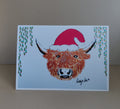 Printed artwork festive Christmas Card featuring a Highland Coo wearing a red Santa hat, leafy foliage hangs down on each side of the coo. Original artwork & printed by Cathy's Art*