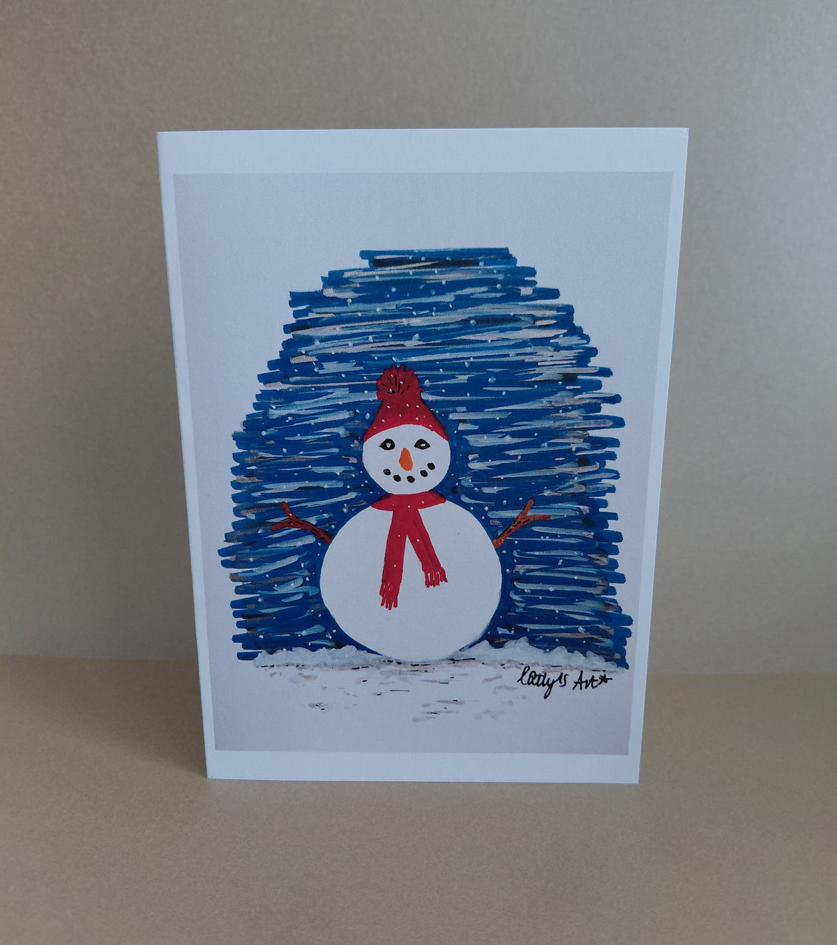 A printed artwork Christmas card featuring a snowman with stick arms, wearing a red bobble hat and red scarf. Behind is a blue snowy night sky. Original artwork & printed by Cathy's Art*