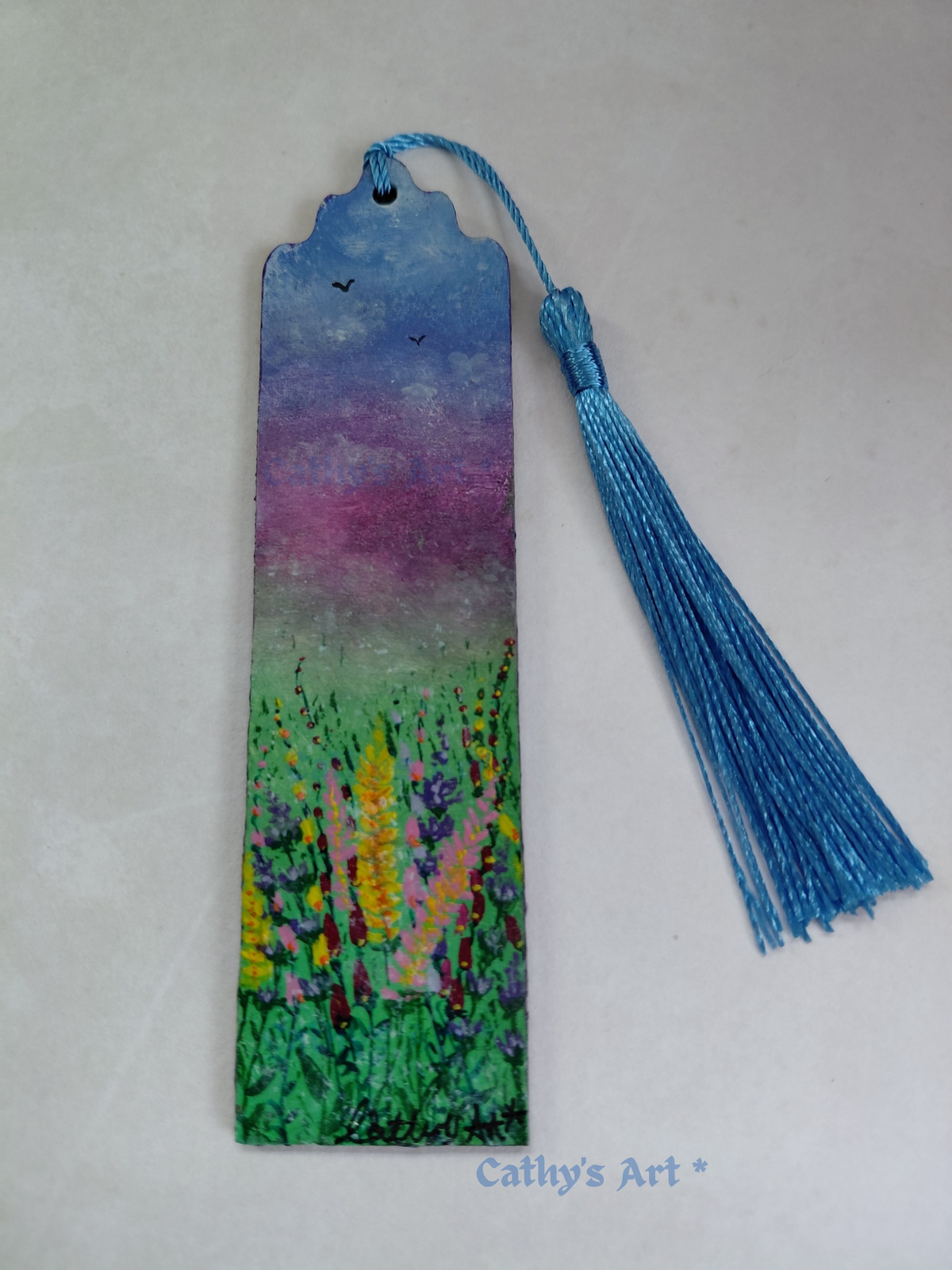 A hand painted bookmark painted with a colourful floral landscape scene, featuring a blue and purple sky with birds and a green field with wild flowers. It has a blue tassel and is photographed on a light background with a 'Cathy's Art *' watermark at the bottom. Original painted wooden bookmark by Cathy's Art*