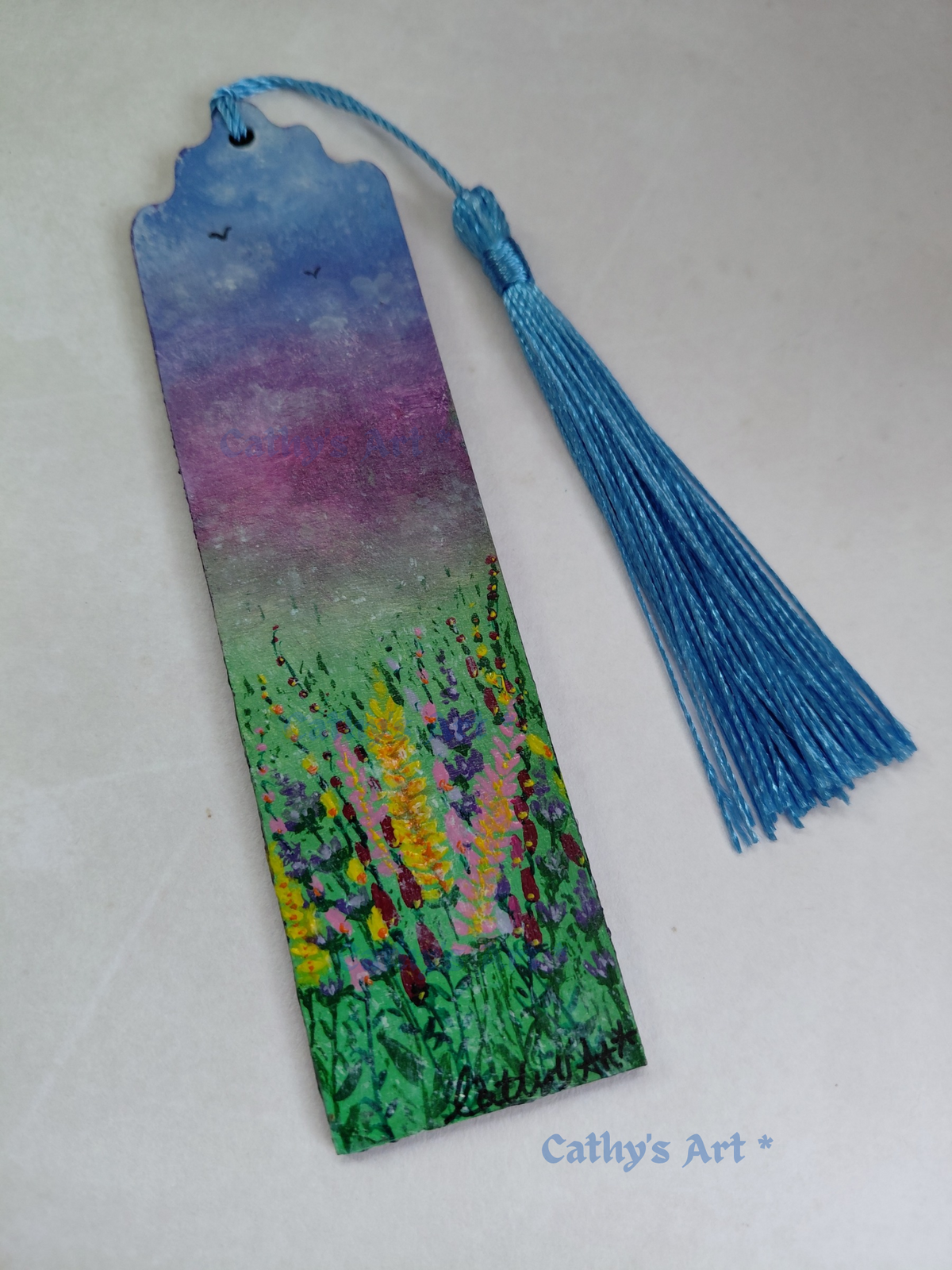 A hand painted bookmark painted with a colourful floral landscape scene, featuring a blue and purple sky with birds and a green field with wild flowers. It has a blue tassel and is photographed on a light background with a 'Cathy's Art *' watermark at the bottom. Original painted wooden bookmark by Cathy's Art*