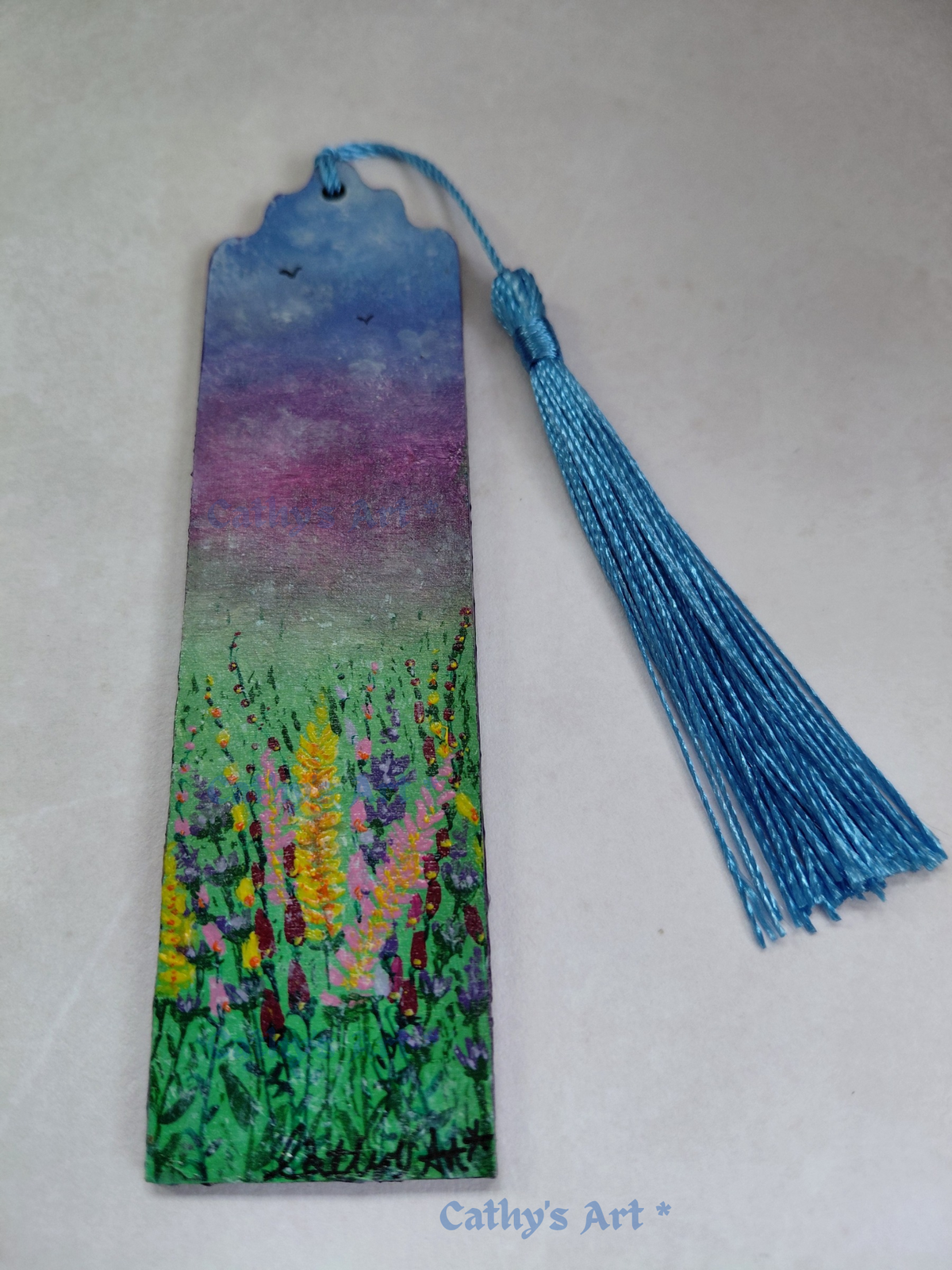 A rectangular wooden bookmark with a shaped top, painted with a colourful landscape scene of wildflowers under a blue and purple sky, with birds, featuring a blue tassel and the text 'Cathy's Art' printed at the bottom. Original painting on wooden bookmark by Cathy's Art*