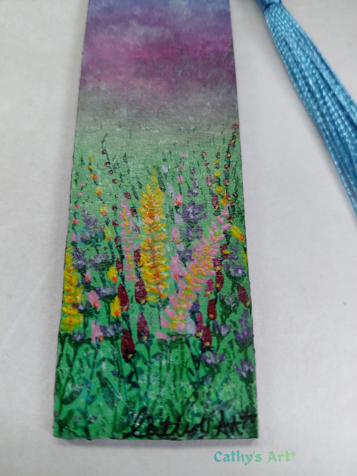 A hand painted bookmark featuring a wildflower meadow scene in green, purple, pink, red and yellow tones, with a blue tassel attached at the top right. The bookmark is photographed on a light neutral background, and the text 'Cathy's Art*' appears in the lower right corner. Original hand painted wooden bookmark by Cathy's Art*