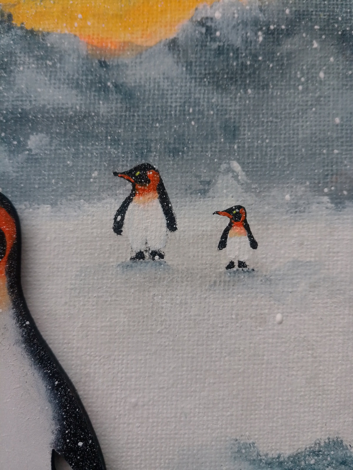 A close-up of a painted canvas showing two penguins standing in snowy white ground under grey mountains with a yellow-orange horizon. Part of a large penguin is visible along the left edge. Original acrylic canvas painting by Cathy's Art*