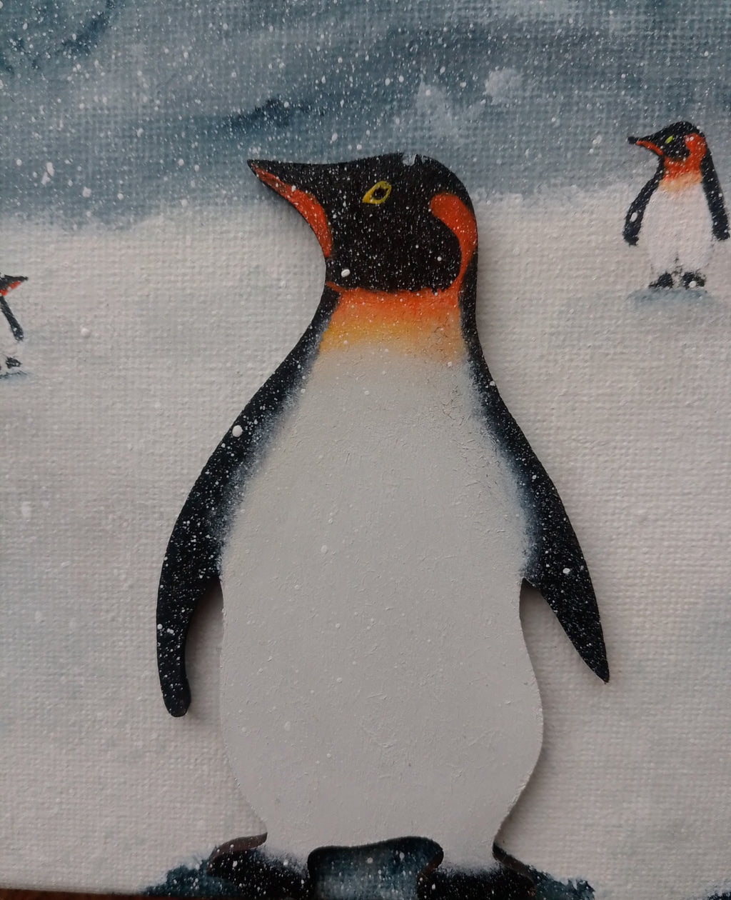 A close-up of a hand-painted canvas featuring a penguin in a snowy winter scene, with other penguin figures in the background. Colours of white, black, orange and grey. original acrylic canvas painting by Cathy's Art*