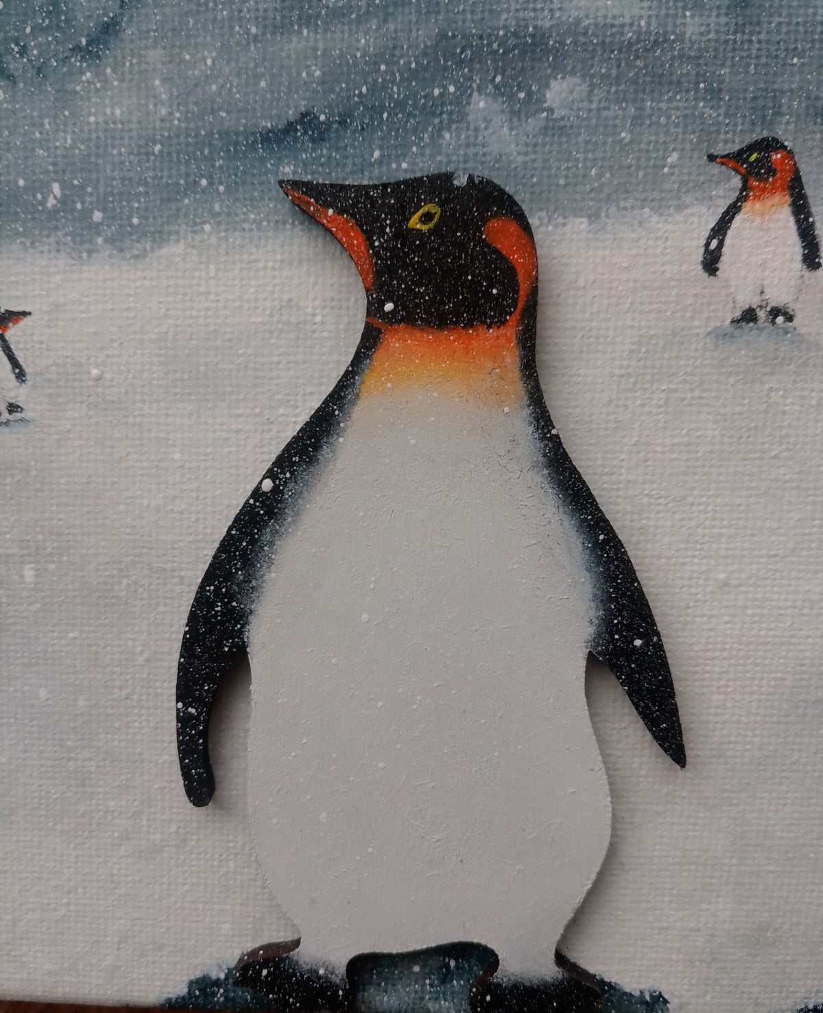 A close-up of a hand-painted canvas featuring a penguin in a snowy winter scene, with other penguin figures in the background. Colours of white, black, orange and grey. original acrylic canvas painting by Cathy's Art*
