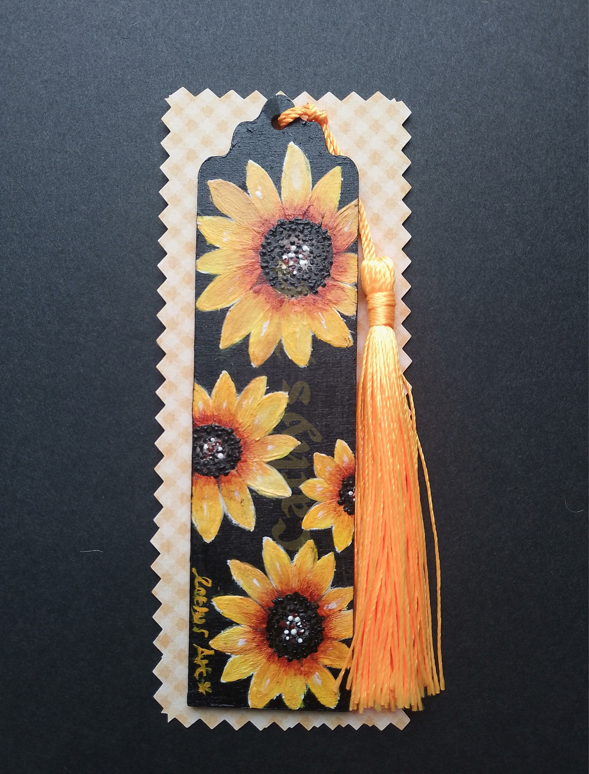 A decorative hand painted bookmark featuring a black background with large yellow sunflower, on white and yellow checked packaging on a black background, and a long orange tassel on the right side. Original hand painted bookmark by Cathy's Art*