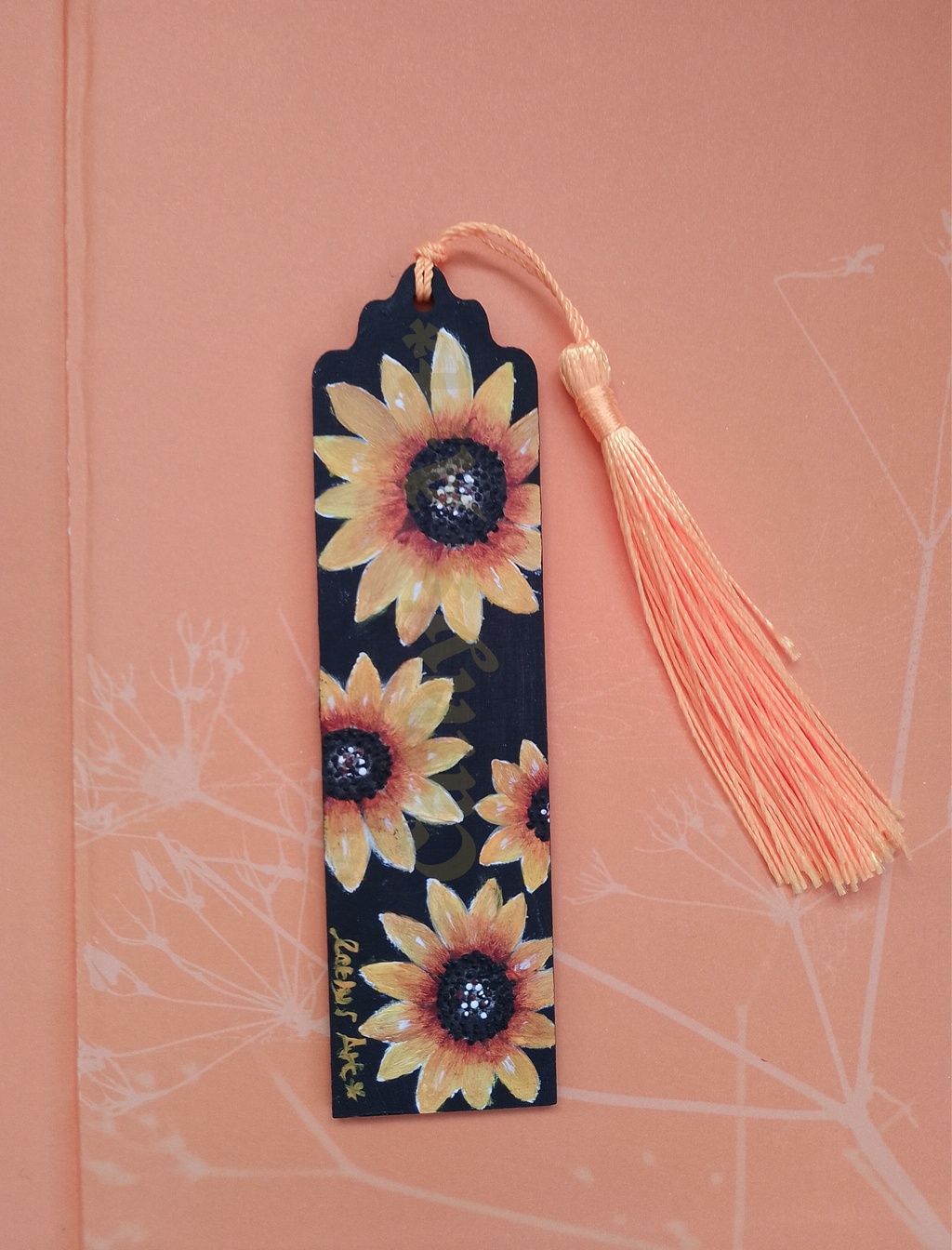 A decorative bookmark with a black background and hand painted sunflower pattern, featuring four sunflowers and a yellow tassel, placed on a pale orange peach-coloured pattern background. Original hand painted bookmark by Cathy's art*
