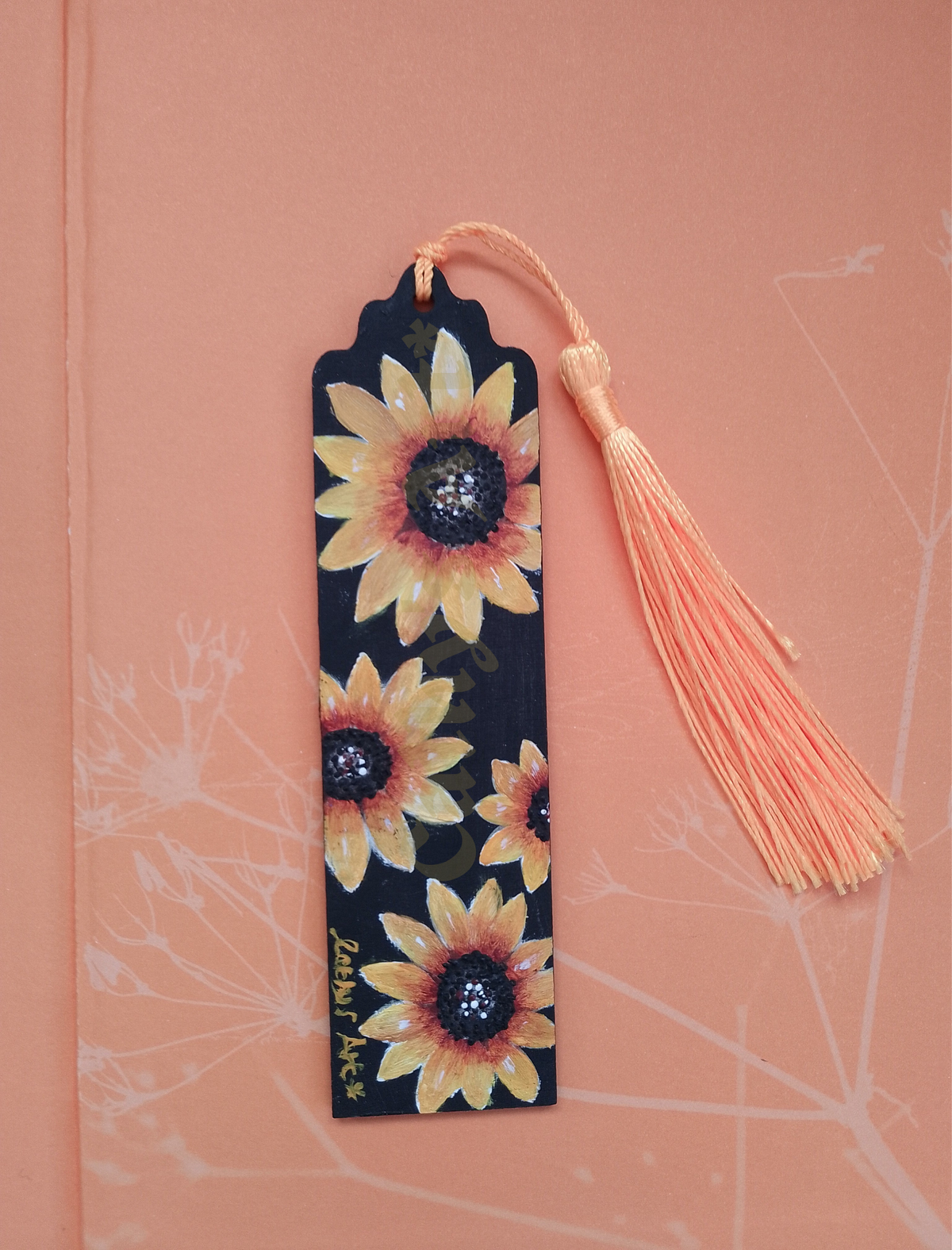 A decorative bookmark with a black background and hand painted sunflower pattern, featuring four sunflowers and a yellow tassel, placed on a pale orange peach-coloured pattern background. Original hand painted bookmark by Cathy's art*