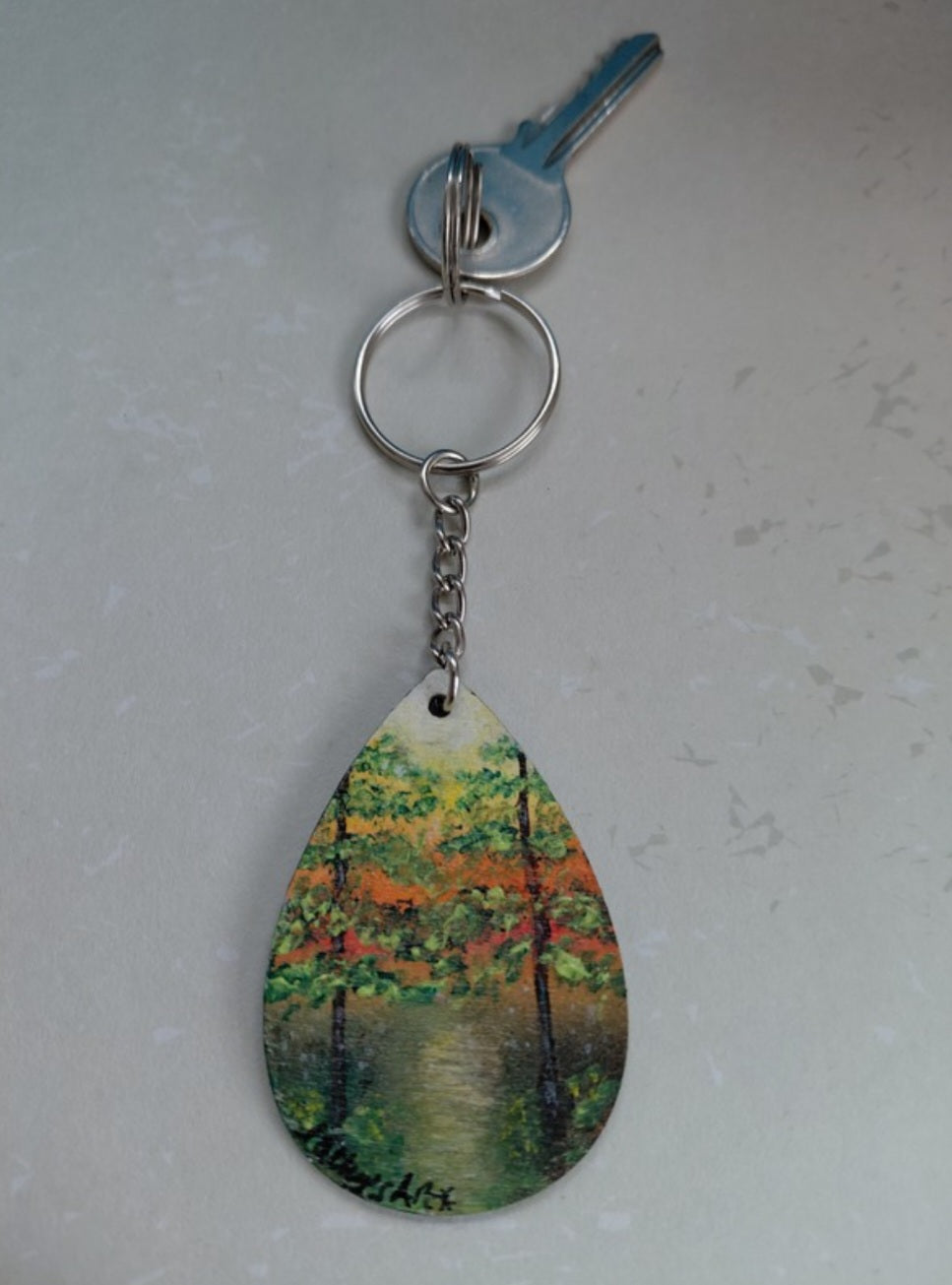 A teardrop-shaped keychain featuring a hand painted landscape  of a tree-lined path with green, and yellow foliage, with an orange and yellow sky, attached to a silver metal keyring with a key on a light grey textured background. Original painting by Cathy's Art*