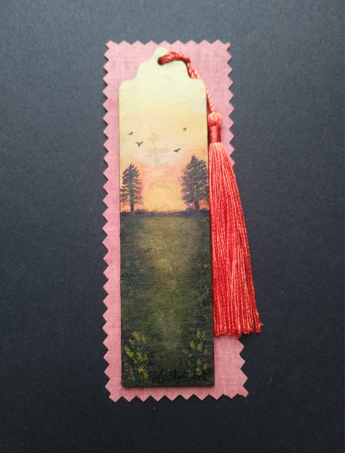 A hand painted bookmark, featuring a  landscape scene of a green field under a sunset sky with trees and birds. An orange tassel is attached at the top. the bookmark is on pale orange packaging on a black background. By Cathy's Art* 