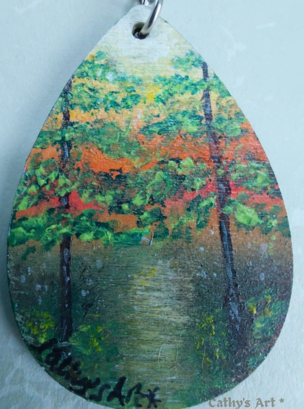 A teardrop-shaped wooden keychain featuring a painted forest scene with a yellow orange sunset sky, hanging from a silver metal ring against a light grey textured background. Original painting by Cathy's Art*