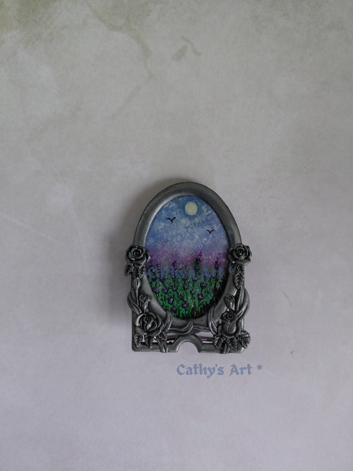 An ornate oval metal picture frame with floral rose details on the sides, holding a miniature landscape painting of a lavender field under a blue sky with the sun, displayed on a textured lilac background with a watermark 'Cathy's Art *'. Original miniature framed painting by Cathy's Art*