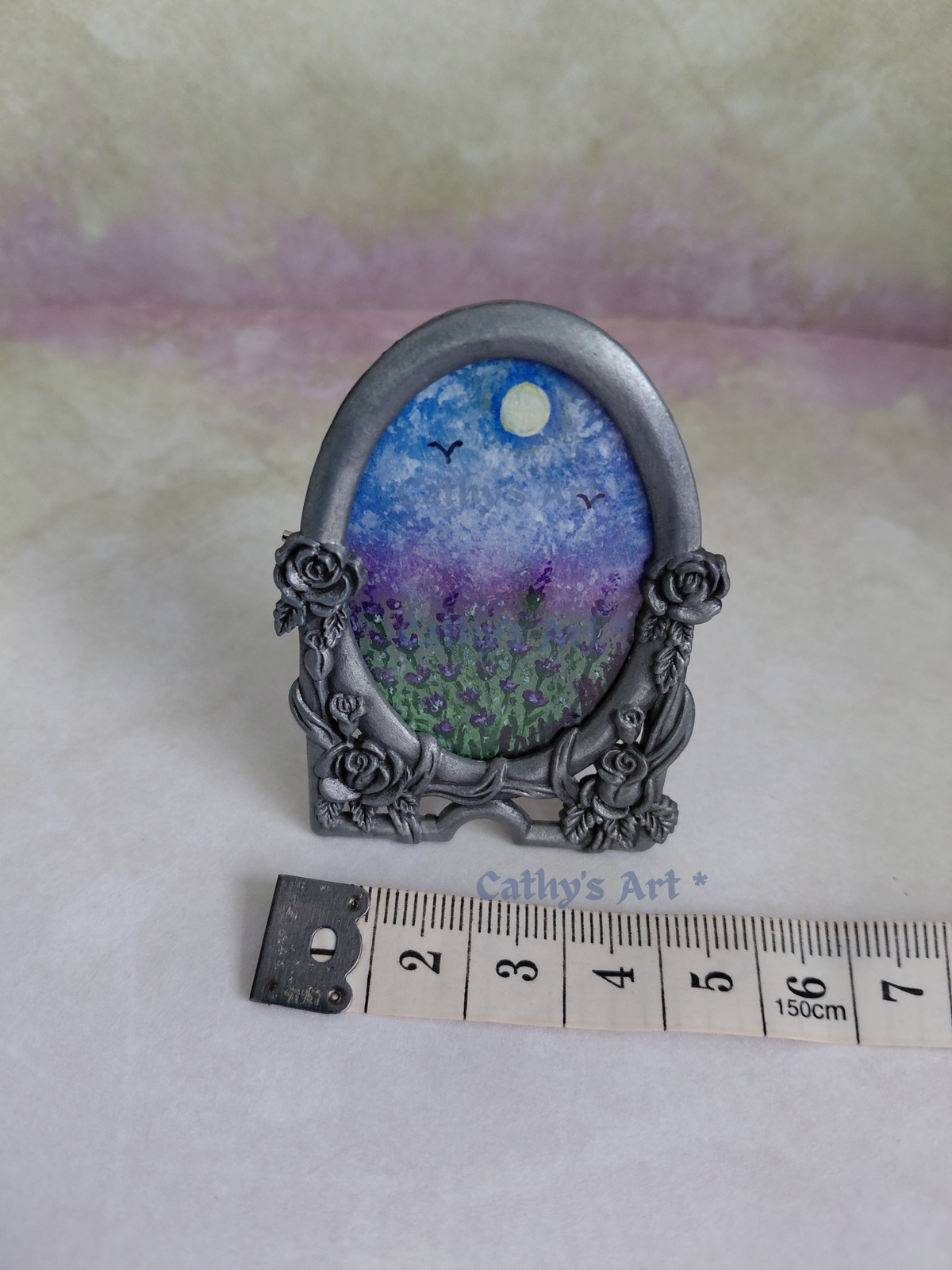 An oval miniature art piece in an ornate silver frame, featuring a blue sky with a sun, birds, and a purple field of lavender. A tape measure is placed below the frame to show scale against a textured lilac background. Original framed miniature painting by Cathy's Art*