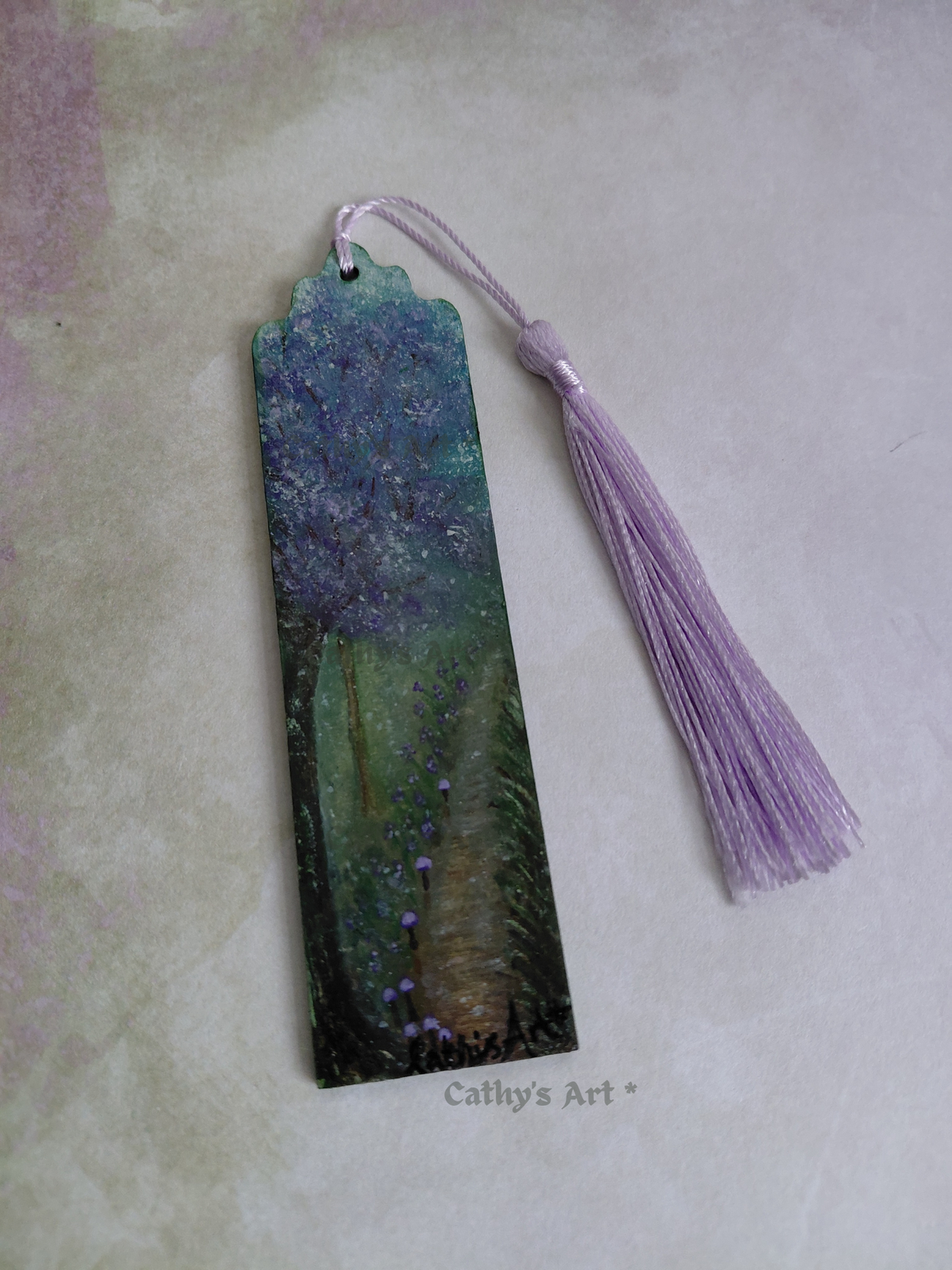 A hand painted enchanted bookmark with a purple tree, tiny purple mushroom line a pathway, with a lilac tassel. photographed on a lilac and green background. Original hand painted bookmark by Cathy's Art*