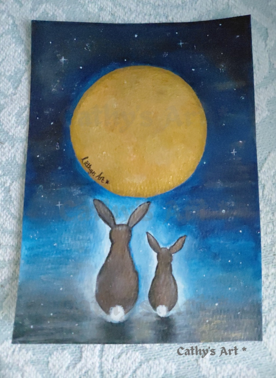 An original painting showing two brown hares facing a large glowing yellow moon against a dark blue starry night sky, placed on a light textured fabric background. The artist signature 'Cathy's Art' is visible near the bottom right of the artwork. Original whimsical painting by Cathy's Art*