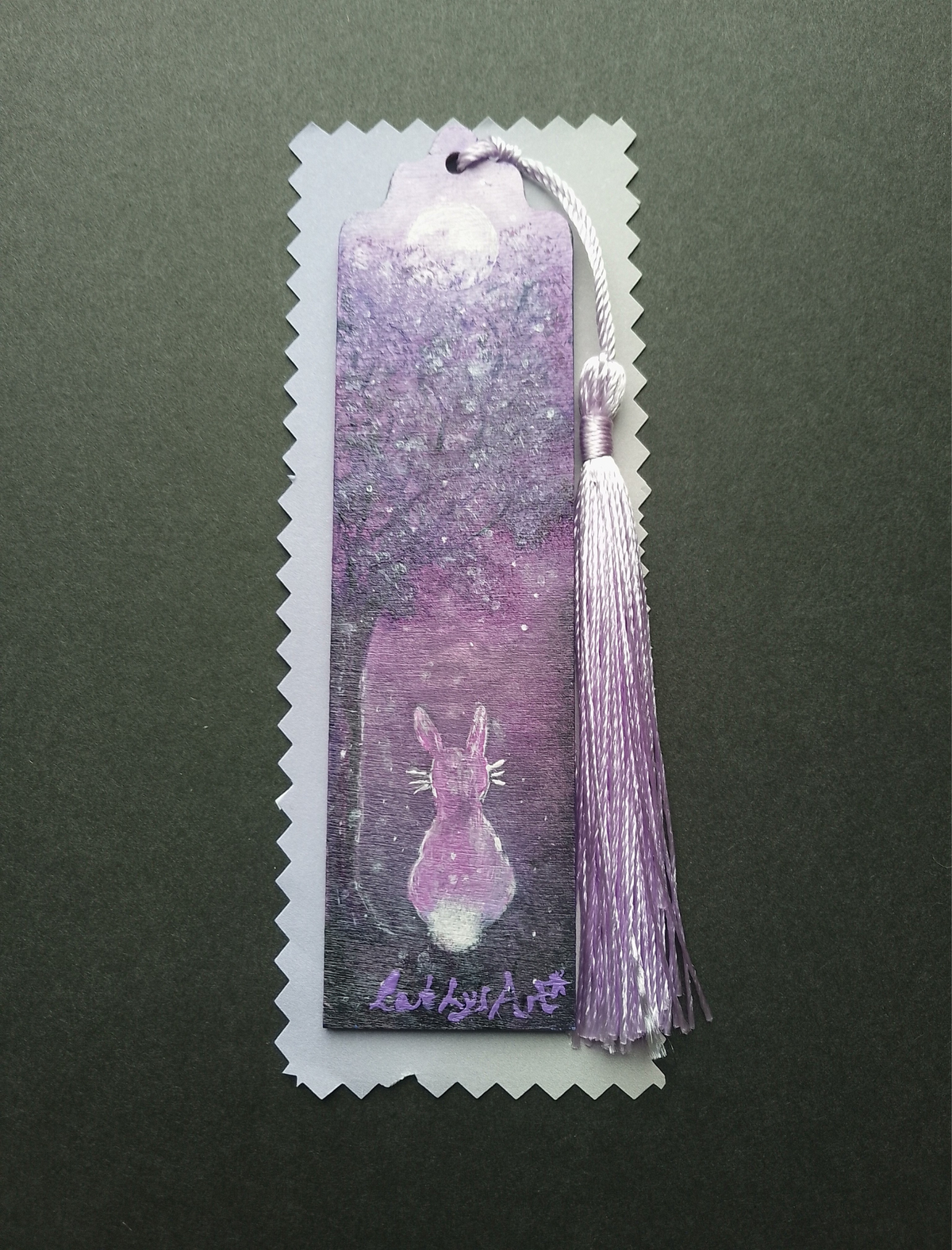 A hand painted wooden bookmark with a tassel, featuring a purple forest illustration with a hare and moon, on lilac packaging shown against a black background. By Cathy's Art*