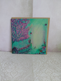 A square art print greeting card featuring a whimsical landscape painting with a pale lemon and green background, pink flowering blossom tree, an gateway, butterflies, a small rabbit and flowers, displayed against a textured light wall on a patterned surface. Original printed artwork greeting card by Cathy's Art*