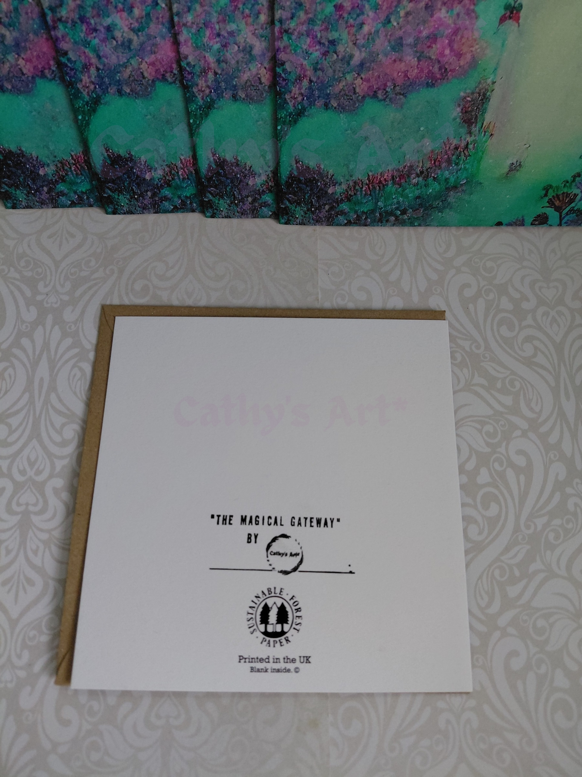 A printed greeting card titled "The Magical Gateway" by Cathy's Art*, placed on a patterned white surface with a kraft envelope behind it and printed greeting cards above it. Original printed artwork greeting card by Cathy's Art