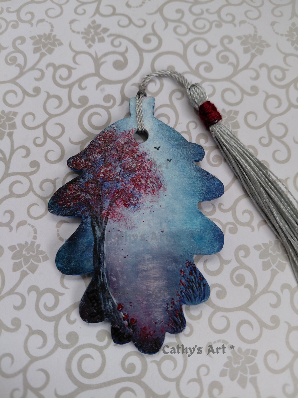 A hand-painted wooden bookmark shaped like an acorn leaf with a blue and white moonlit sky and path, a grey and red tree, birds in the sky, red flowers and tiny red mushrooms, with a silver tassel. displayed on a light patterned background. The text 'Cathy's Art*' is visible on the image. Original hand painted wooden acorn leaf bookmark by Cathy's Art*