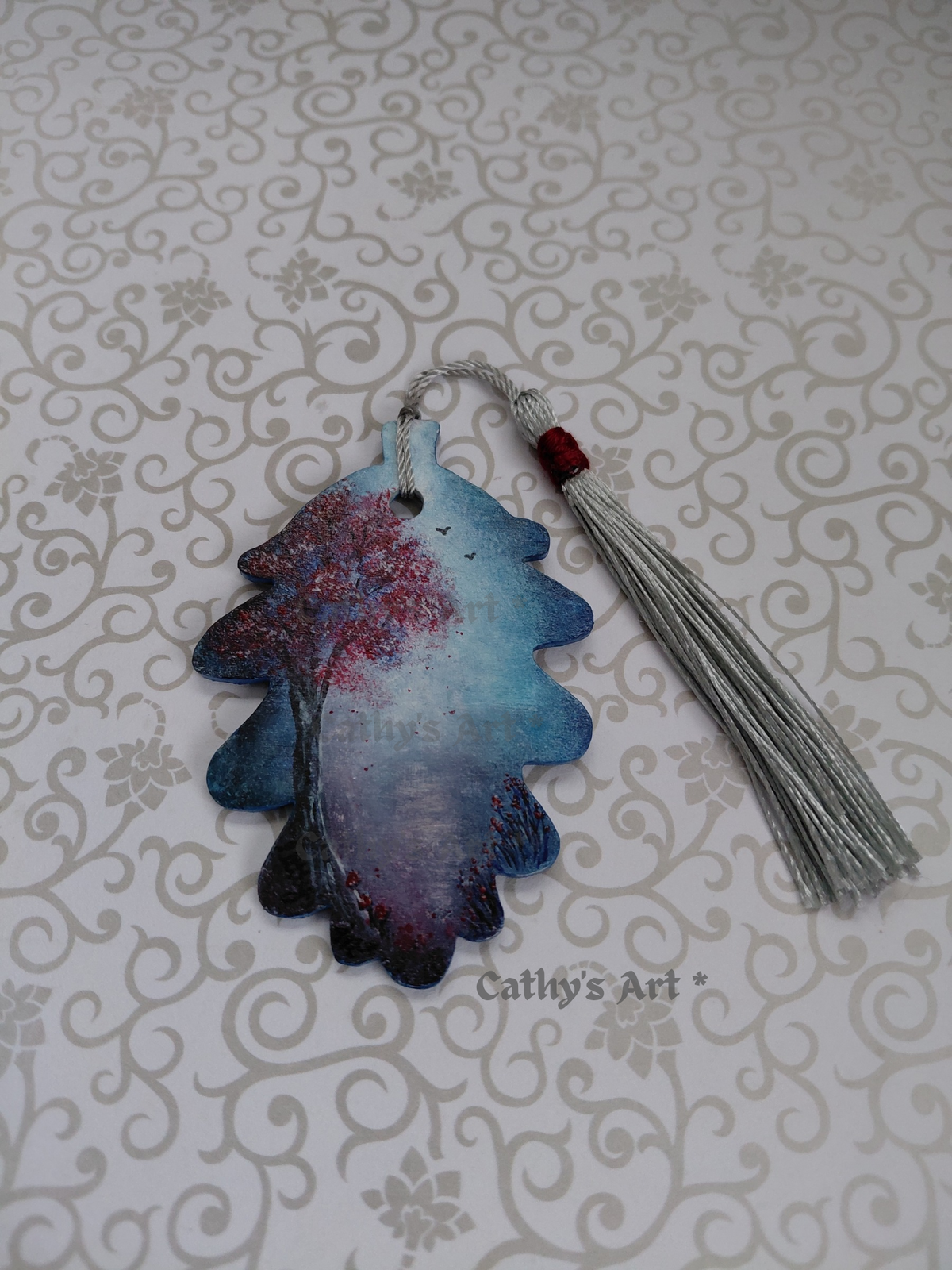 A hand-painted wooden bookmark shaped like an acorn leaf with a blue and white moonlit sky and path, a grey and red tree, birds in the sky, red flowers and tiny red mushrooms, with a silver tassel. displayed on a light patterned background. The text 'Cathy's Art*' is visible on the image. Original hand painted wooden acorn leaf bookmark by Cathy's Art*