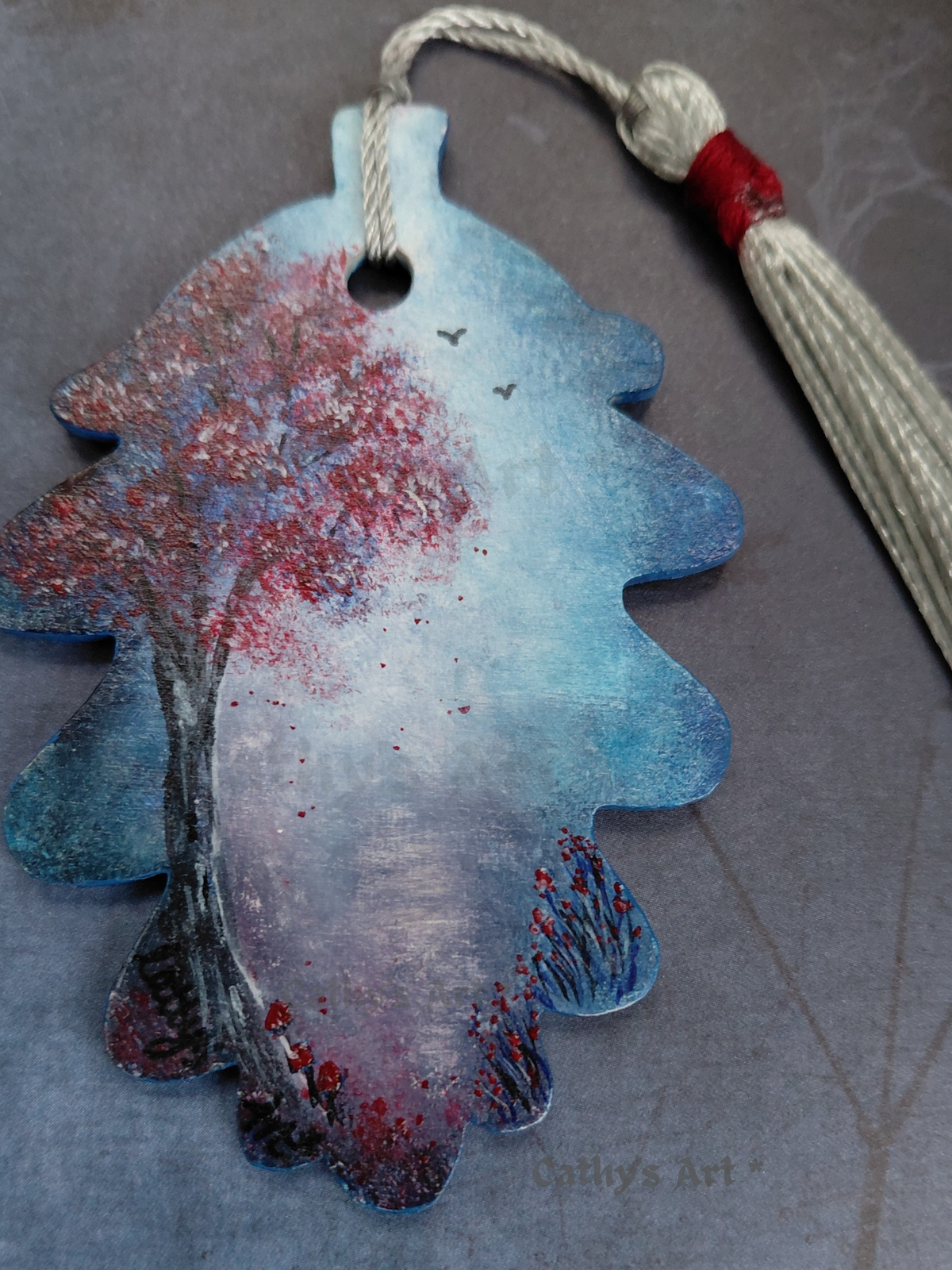 A close up painting of a hand painted acorn leaf shaped bookmark. Red and grey tree, blue and white moonlit background with a moonlit path, red flowers and mushrooms, and birds in the sky. Photographed on a dark grey background. Original hand painted bookmark by Cathy's Art*