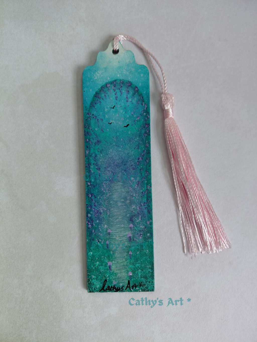 A hand painted bookmark with a pink tassel, featuring a turquoise, green and purple land with birds, a mushroom lined path and an archway with cascading purple foliage, photographed on a textured light background. Original hand painted bookmark by Cathy's Art*