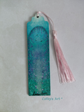 A hand painted bookmark with a pink tassel, featuring a turquoise, green and purple land with birds, a mushroom lined path and an archway with cascading purple foliage, photographed on a textured light background. Original hand painted bookmark by Cathy's Art*