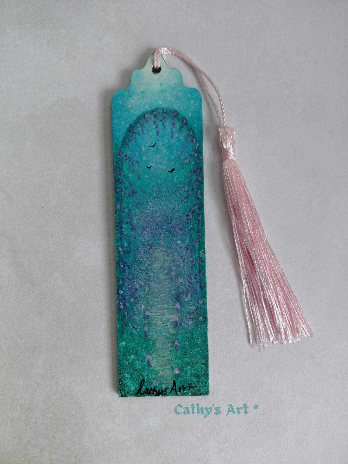 A hand painted bookmark with a pink tassel, featuring a turquoise, green and purple land with birds, a mushroom lined path and an archway with cascading purple foliage, photographed on a textured light background. Original hand painted bookmark by Cathy's Art*