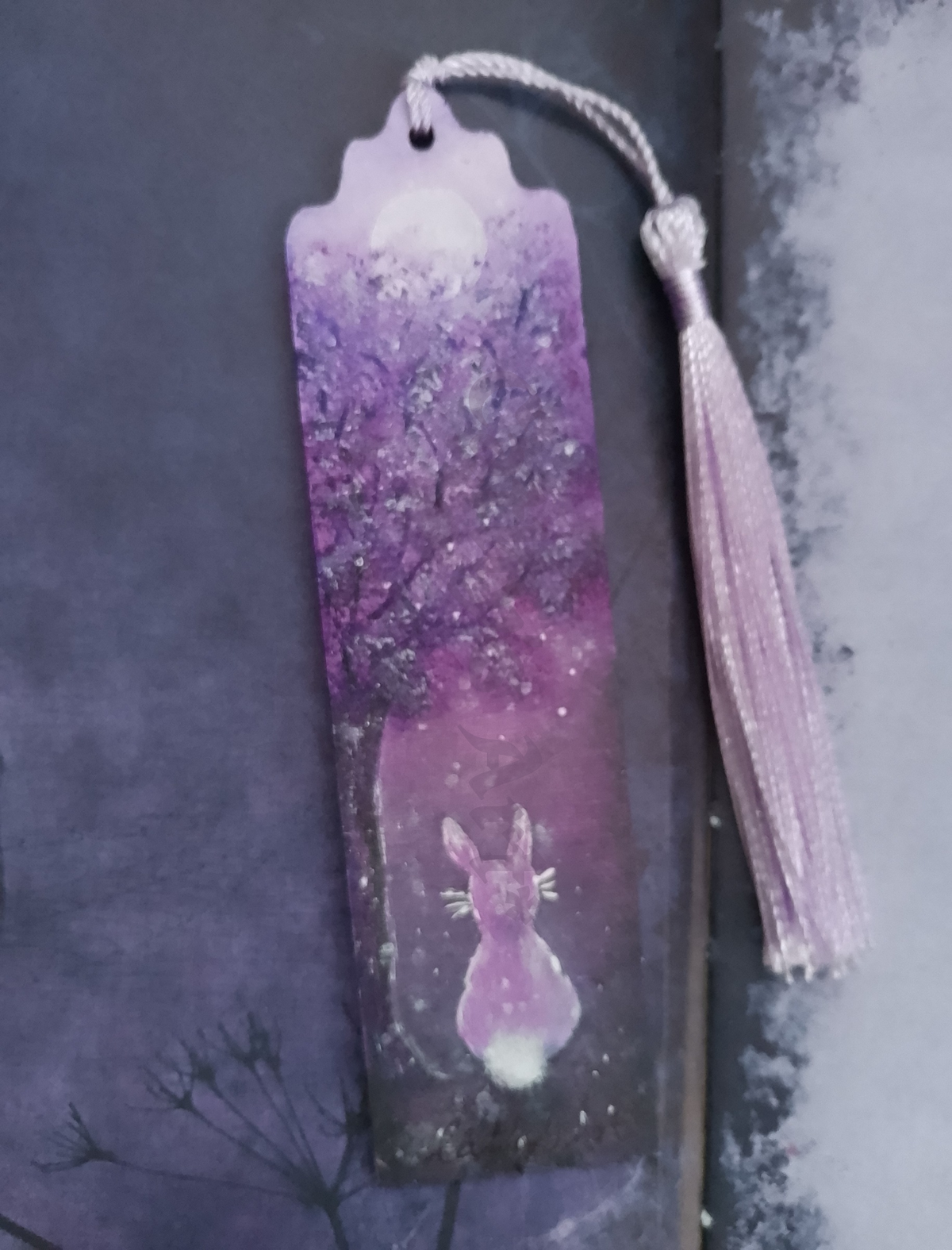 A hand painted wooden bookmark with a tassel, featuring a purple forest illustration with a hare and moon, shown against a dark textured background. By Cathy's Art*
