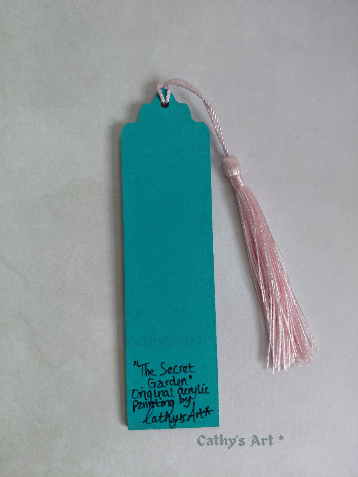 The back view of a hand painted bookmark, in turquoise colour, featuring hand written text "The Secret Garden" Original acrylic painting by Cathy's Art* On a light coloured background. original hand painted bookmark by Cathy's Art*