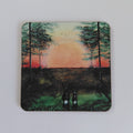 A square coaster with rounded corners featuring a painted landscape scene of two hares facing a large orange sunset between trees, shown on a plain white background. A print of Tranquil sunset, original artwork by Cathy's Art* on a cork backed coaster.