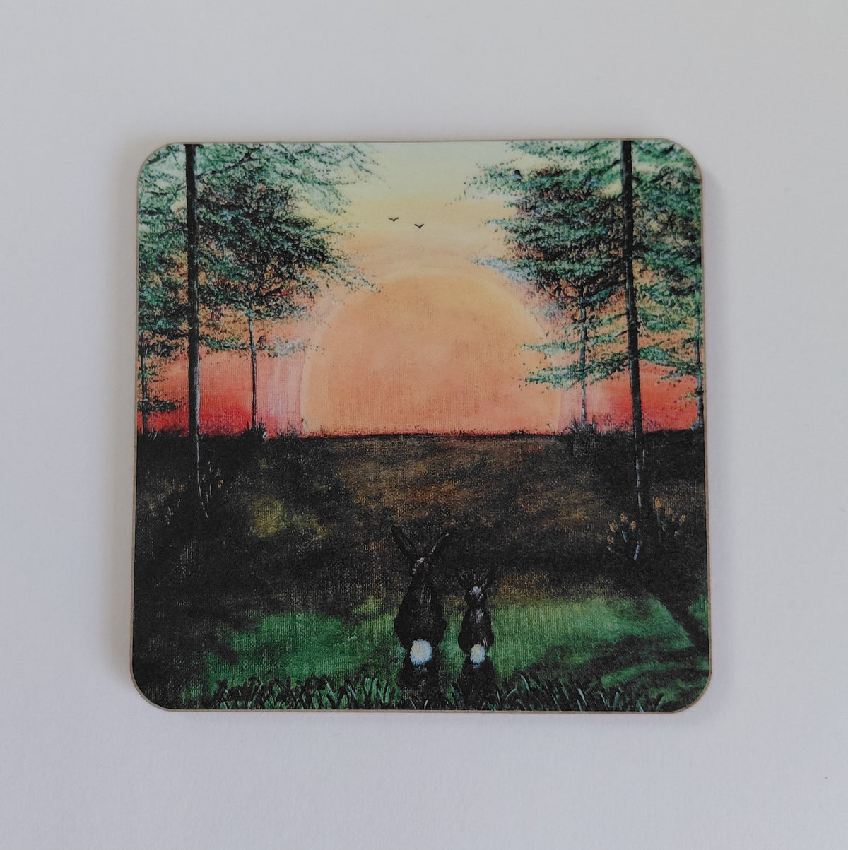 A square coaster with rounded corners featuring a painted landscape scene of two hares facing a large orange sunset between trees, shown on a plain white background. A print of Tranquil sunset, original artwork by Cathy's Art* on a cork backed coaster.