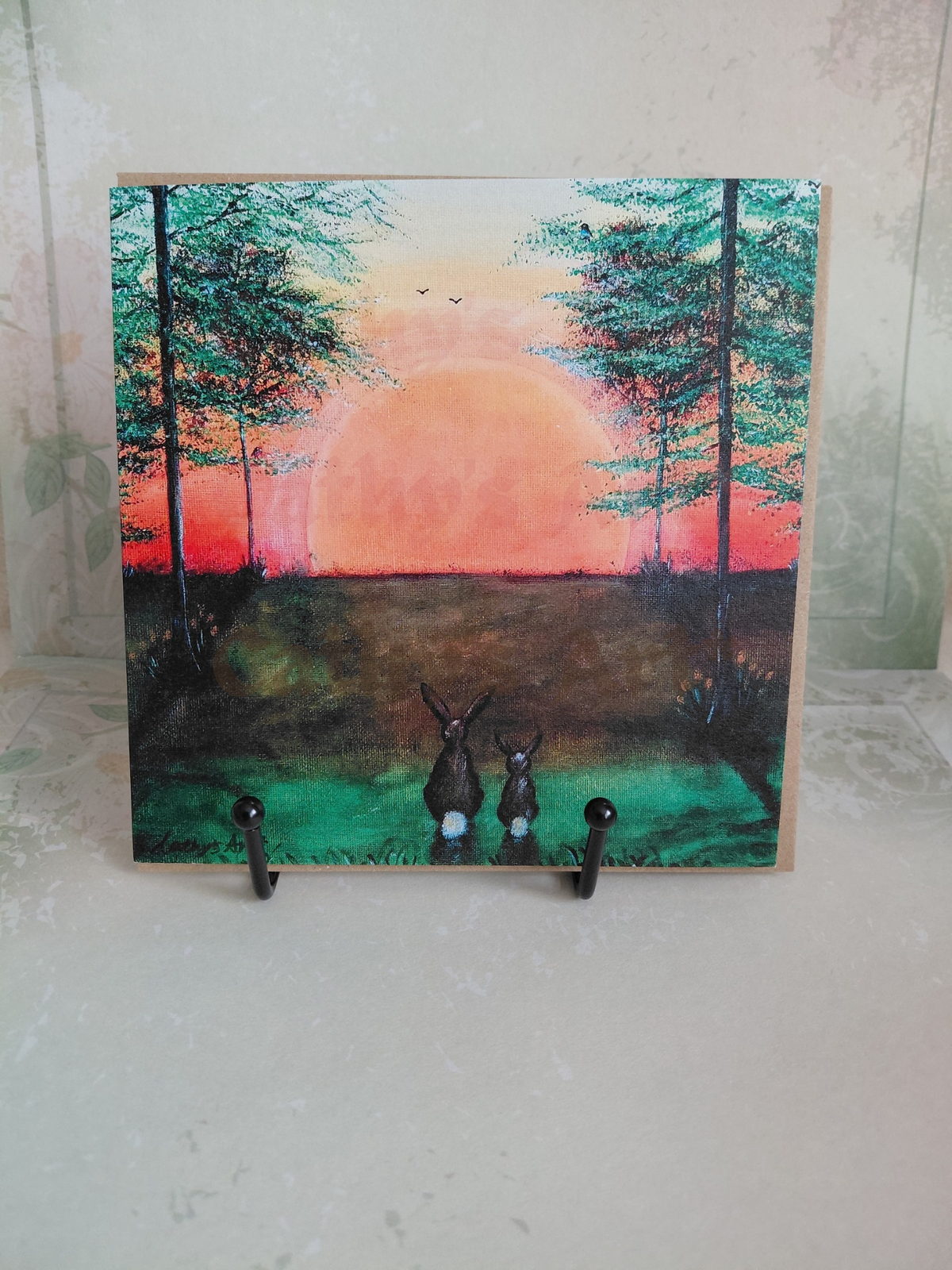 A square artwork printed greeting card featuring a painted forest landscape at sunset with two hares in the foreground, displayed on a light surface with two small black hooks supporting it. Original printed artwork greeting card by Cathy's Art*