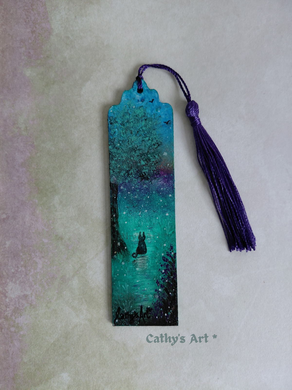 A hand painted whimsical bookmark in turquoise, purple, black and green colours. A black cat sits under a tree, birds fly in the sky, purple flowers bloom, magical orbs dance in the air. With purple tassel, on a textured pale lilac and green . Original hand painted bookmark by Cathy's Art*