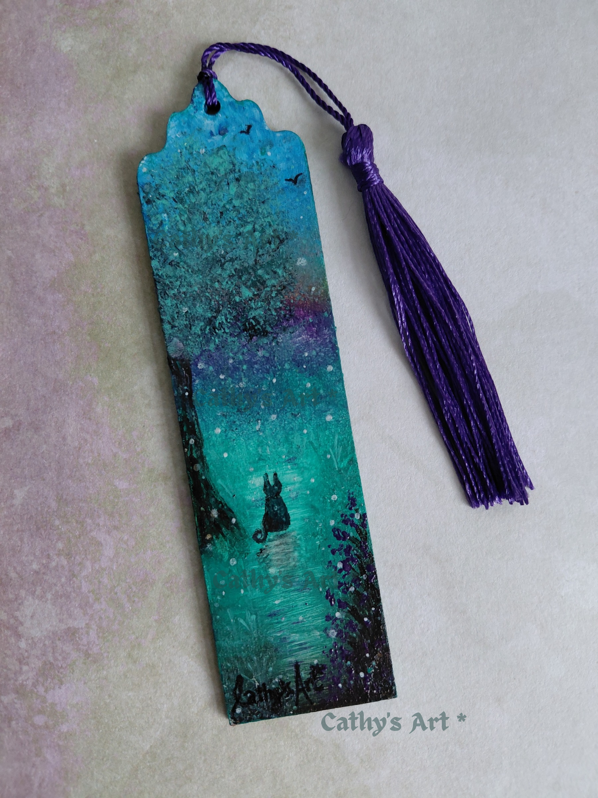 A hand painted whimsical bookmark in turquoise, purple, black and green colours. A black cat sits under a tree, birds fly in the sky, purple flowers bloom, magical orbs dance in the air. With purple tassel, on a textured pale lilac and green . Original hand painted bookmark by Cathy's Art*
