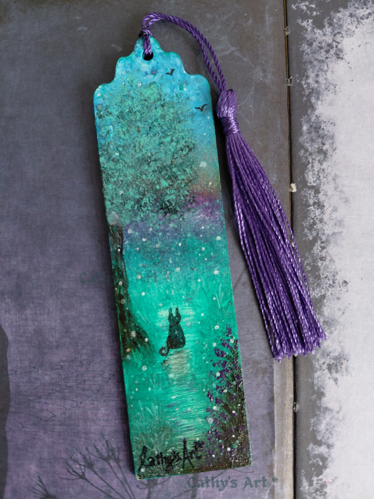 A hand painted whimsical bookmark in turquoise, purple, black and green colours. A black cat sits under a tree, birds fly in the sky, purple flowers bloom, magical orbs dance in the air. With purple tassel, on a textured purple background. Original hand painted bookmark by Cathy's Art*