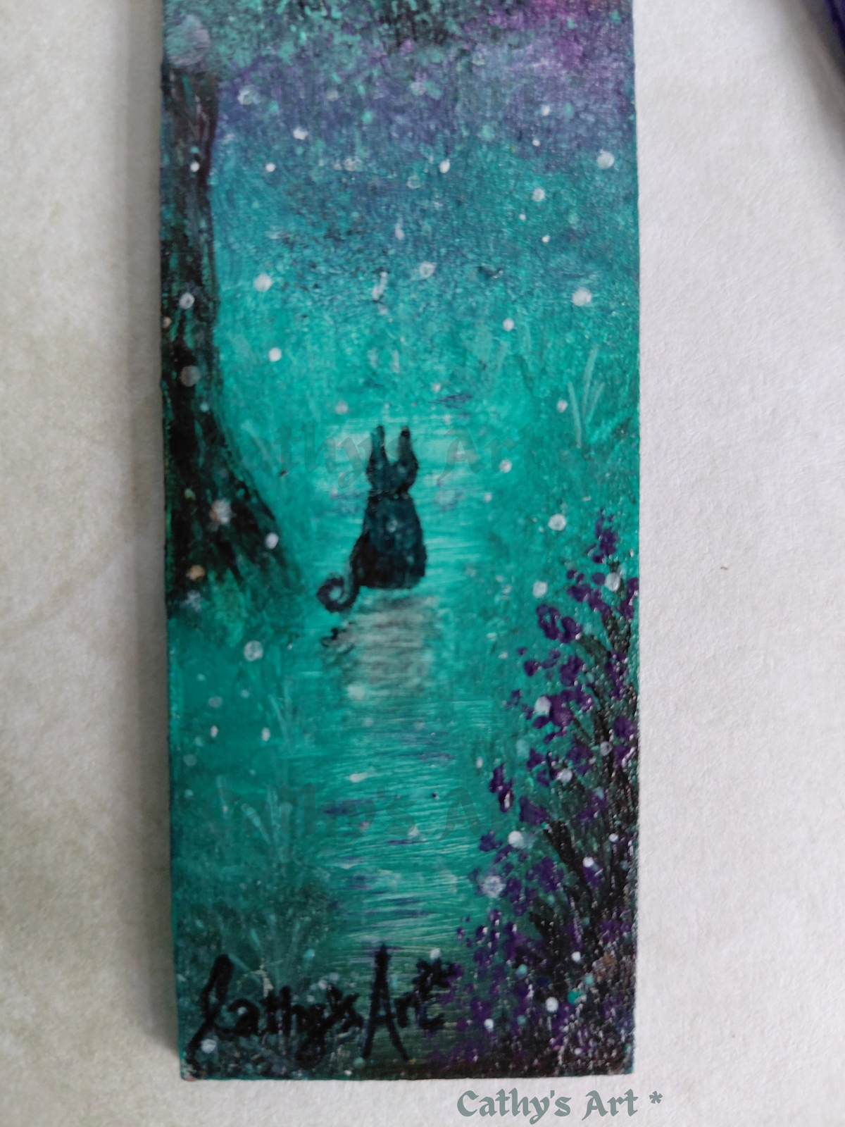 A black cat sits on a path in a magical land, with purple flowers, a tree, and floating orbs. On a pale pink background. Hand painted whimsical bookmark by Cathy's Art*