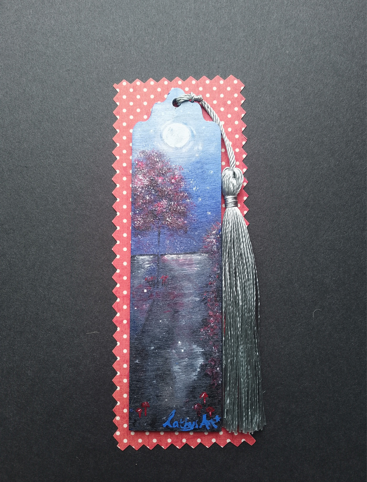 A decorative bookmark with a silver tassel, featuring a hand-painted night landscape scene with a moonlit sky, a red tree, and a reflective path with little red mushrooms, on a red and white spotty packaging on a dark background. Original acrylic painted bookmark by Cathy's Art*