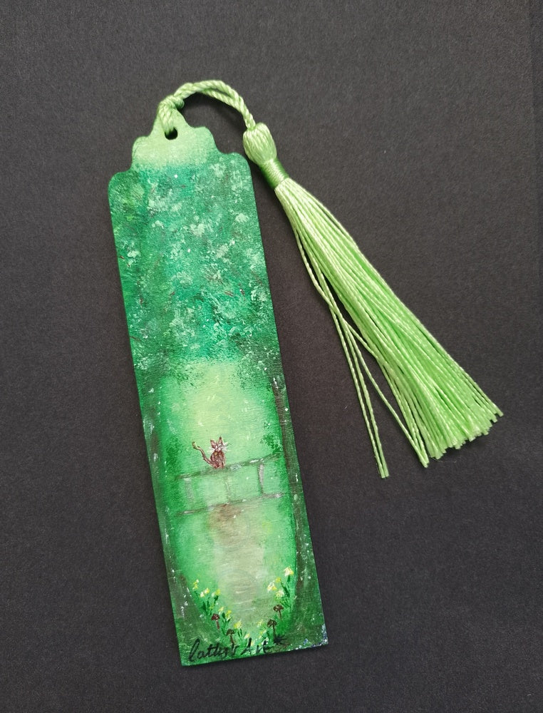A green rectangular bookmark with a green tassel, featuring a whimsical hand painted green tree design with a cat sat on a fence, little yellow flowers and mushrooms grow near the trees, displayed on a black background. Original hand painted bookmark by Cathy's Art*