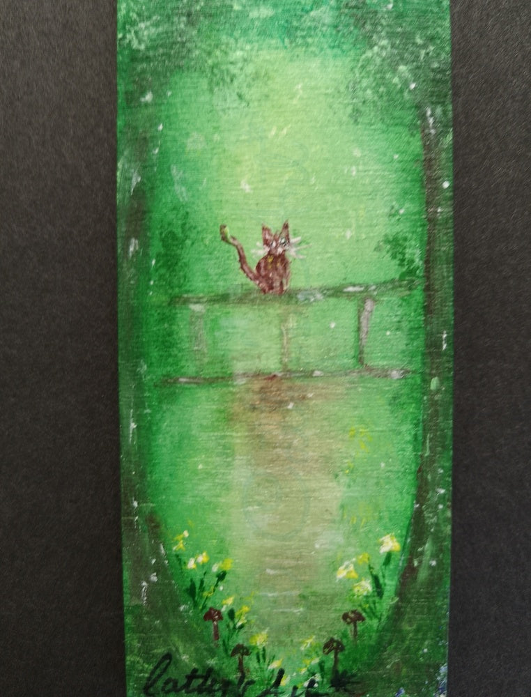 A close up hand painted bookmark in green, brown and yellow tones, featuring trees, a brown cat sat on a fence, and yellow flower and mushrooms. original hand painted bookmark by Cathy's Art*