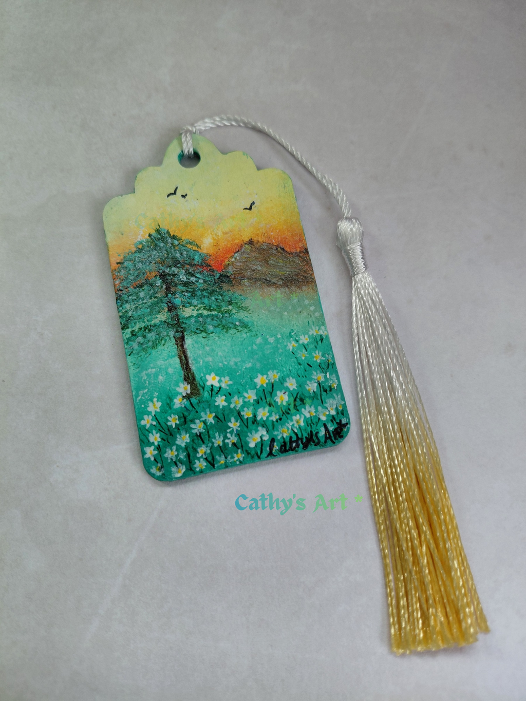 A hand painted bookmark with a peaceful landscape design featuring a tree, white daisy flowers, mountains, and birds under a sunset sky, complete with a white and yellow gold tassel Original painting by Cathy's Art*