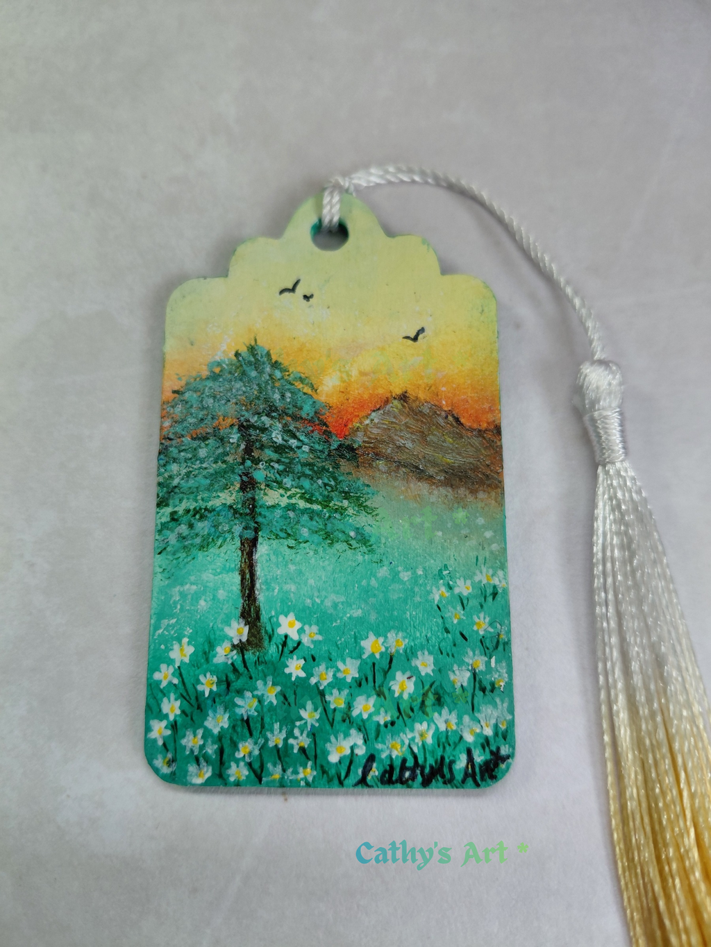 A hand painted bookmark with a peaceful landscape design featuring a tree, white daisy flowers, mountains, and birds under a sunset sky, complete with a white and yellow gold tassel Original painting by Cathy's Art*