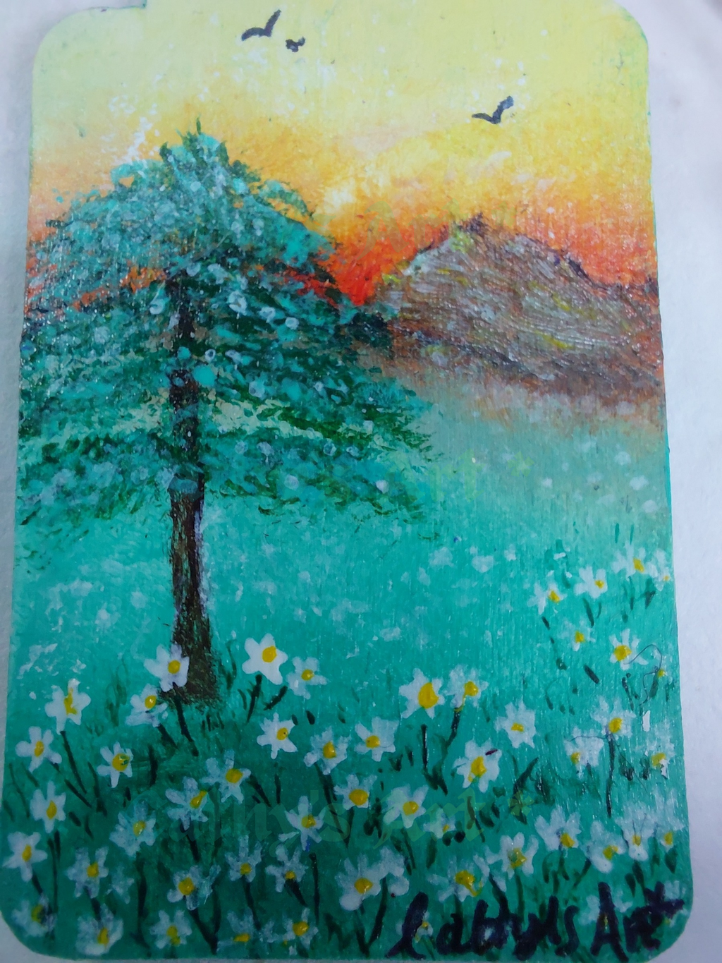 A close up image of a hand painted landscape scene on a wooden bookmark, yellow and orange sunset sky with birds, mountains, a tree and white daises. Hand pained bookmark by Cathy's Art*