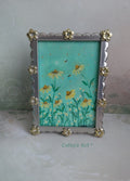 A decorative picture frame with a silver-toned scalloped border and gold-toned floral accents, holding a floral artwork with yellow daisy flowers on a turquoise green background. The frame is photographed against a pale textured wall, with the text 'Cathy's Art' visible near the bottom. Original framed acrylic painting on watercolour paper by Cathy's Art*