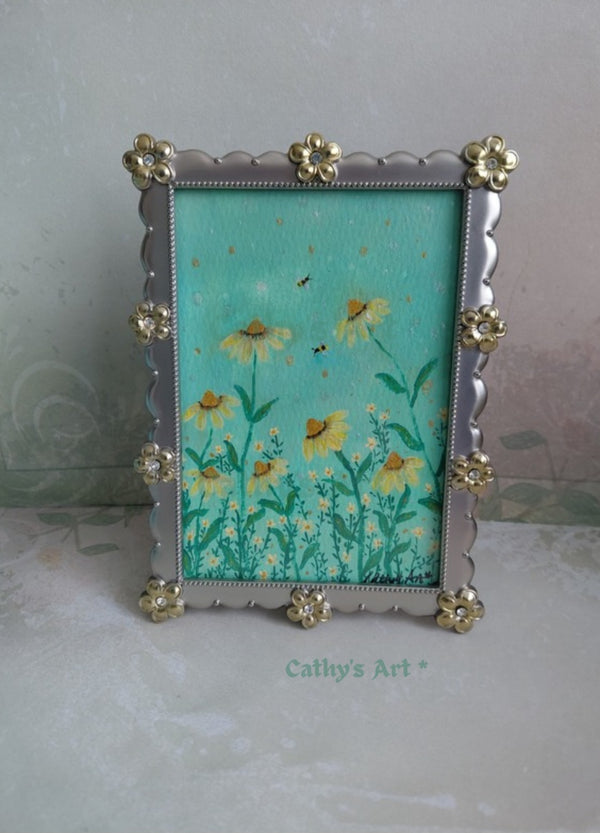 A decorative picture frame with a silver-toned scalloped border and gold-toned floral accents, holding a floral artwork with yellow daisy flowers on a turquoise green background. The frame is photographed against a pale textured wall, with the text 'Cathy's Art' visible near the bottom. Original framed acrylic painting on watercolour paper by Cathy's Art*