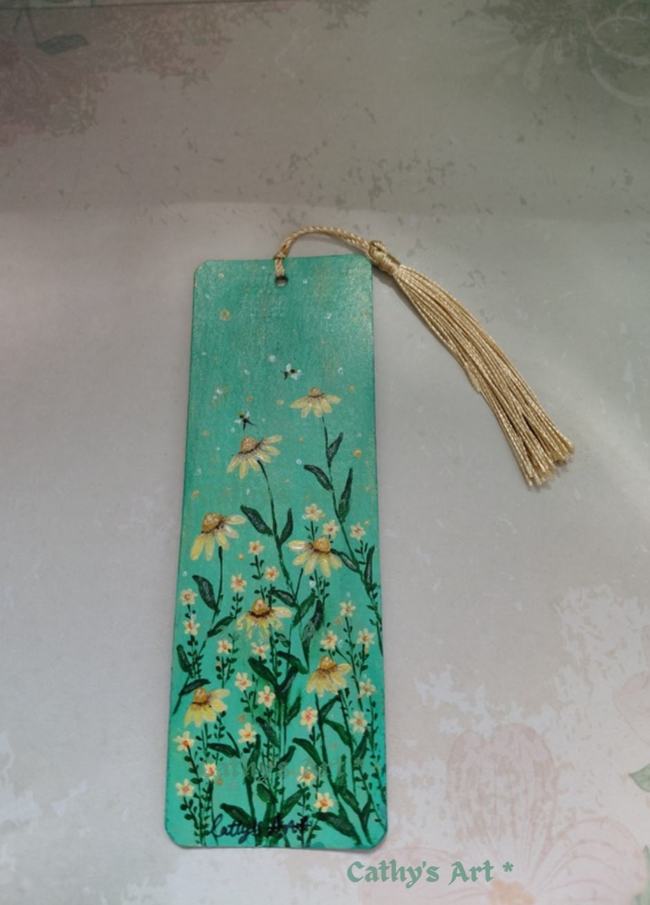 A rectangular pale green painted bookmark with a gold tassel, decorated with hand-painted yellow and white daisies and green stems, with two bees. Photographed on a pale background. Hand painted original bookmark on watercolour paper by Cathy's Art*