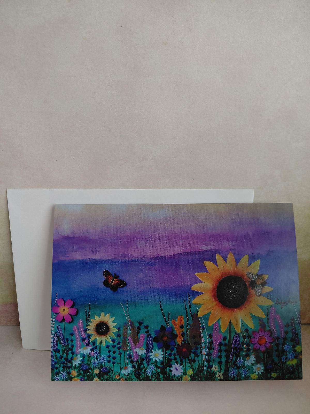 A printed tent style greeting card with a vibrant field of wildflowers with prominent sunflowers, a butterfly, and a bee against a purple sky, with a white envelope displayed on a textured light wall.  A printed greeting card of an original painting by Cathy's art*