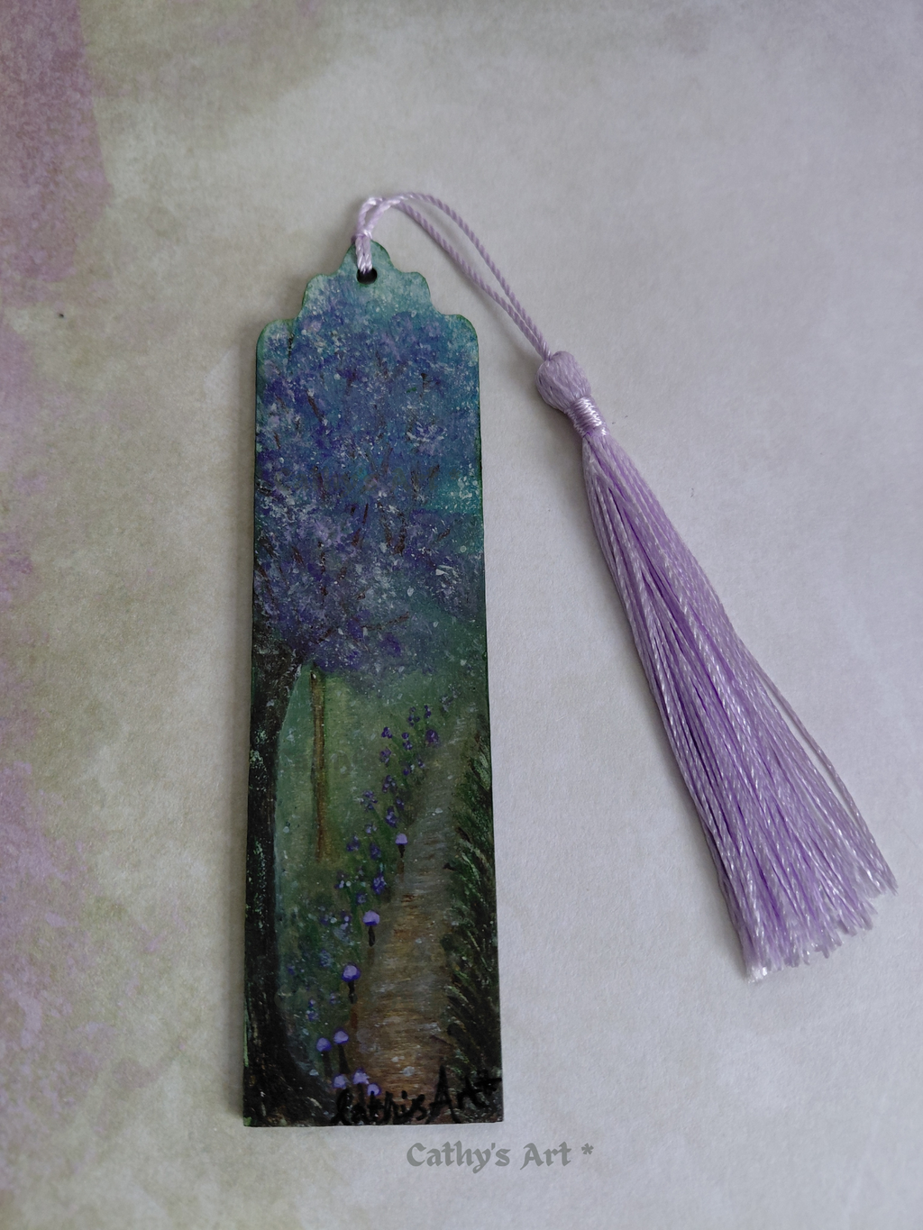A hand painted enchanted bookmark with a purple tree, tiny purple mushroom line a pathway, with a lilac tassel, photographed on a lilac and green background. Original hand painted bookmark by Cathy's Art*