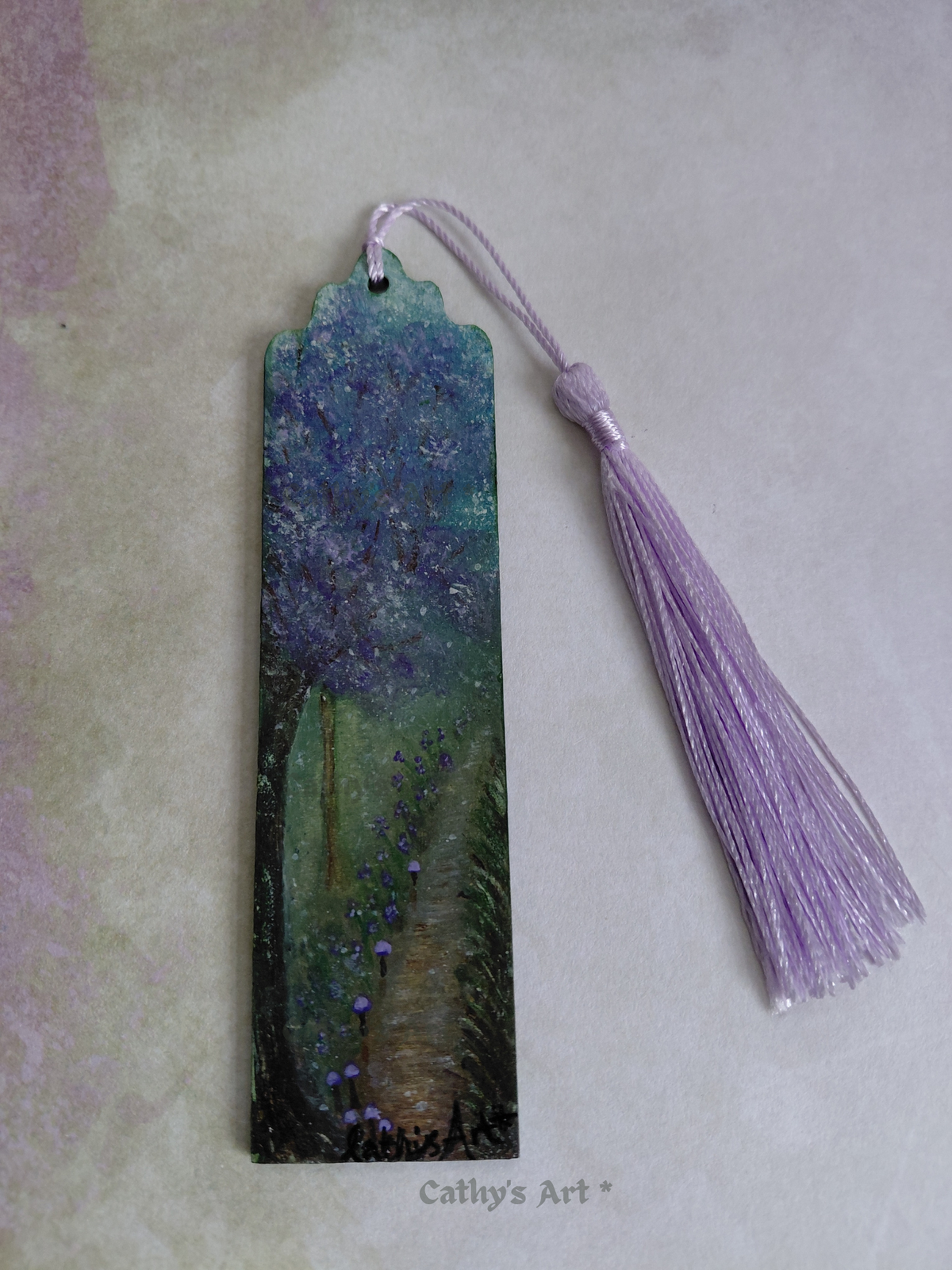 A hand painted enchanted bookmark with a purple tree, tiny purple mushroom line a pathway, with a lilac tassel, photographed on a lilac and green background. Original hand painted bookmark by Cathy's Art*